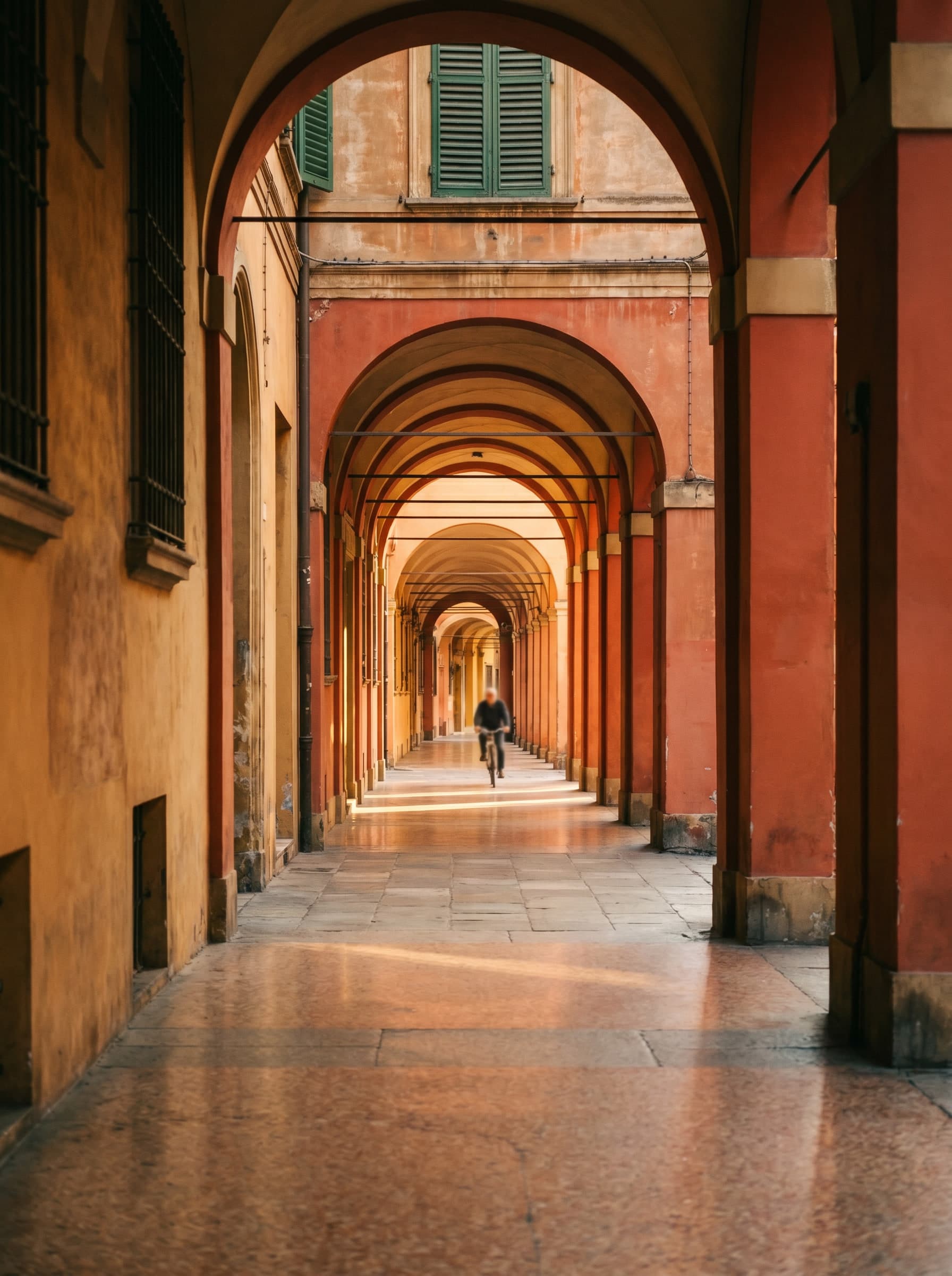 Bologna's iconic terracotta-red porticoes at golden hour, receding arches with a distant cyclist